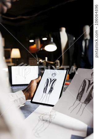 Couturer looking for inspiration for upcoming bespoke comissioned exquisite dress, comparing handdrawn sketch with online outfit. Tailor analyzing designs of fashion attire in tailoring studio 106064896