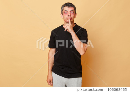 Calm sad man in black T-shirt with bruises and abrasions on his face isolated over beige background standing with finger near lips keeps secret about violence. Calm sad man in black T-shirt with bruises and abrasions on his face isolated over beige background standing with finger near lips keeps secret about violence. 106068465