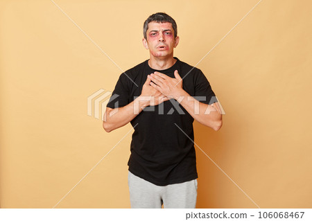 Unhealthy unhappy man in black T-shirt with bruises and abrasions on his face isolated over beige background feeling strong pain chest after car accident. 106068467