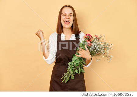 Extremely happy smiling cheerful woman florist wearing brown apron holding bouquet of flowers standing isolated over beige background clenched fists celebrating her business success. Extremely happy smiling cheerful woman florist wearing brown apron holding bouquet of flowers standing isolated over beige background clenched fists celebrating her business success. 106068583