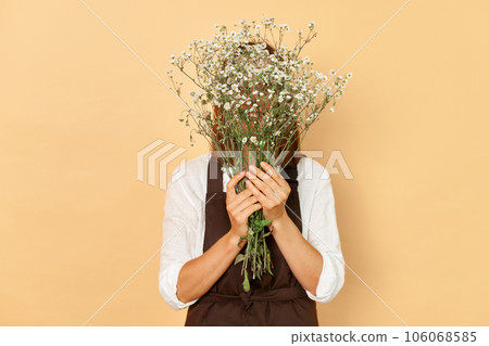 Unknown unrecognizable woman florist wearing brown apron hiding her face behind bouquet of flowers standing isolated over beige background 106068585