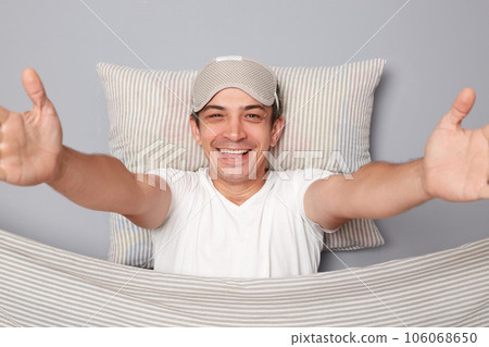 Pleased positive man with resting on white pillow under blanket wearing t-shirt isolated over grey background spread hands showing welcome gesture weekend drowsiness. 106068650