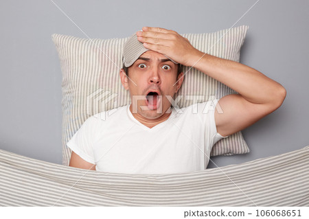 Shocked amazed man with resting on white pillow under blanket wearing t-shirt isolated over grey background looking nat camera with big eyes and open moutn touching head problems Shocked amazed man with resting on white pillow under blanket wearing t-shirt isolated over grey background looking nat camera with big eyes and open moutn touching head problems 106068651
