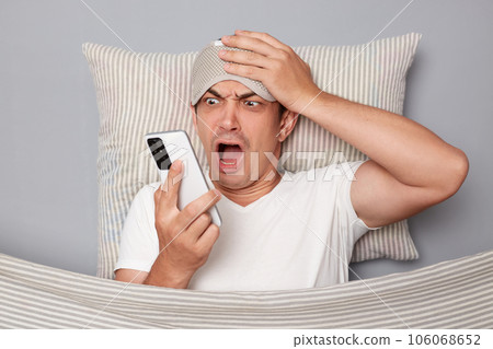 Shocked man with resting on white pillow under blanket wearing t-shirt isolated over grey background waking up looking at smartphone oversleep having despair expression Shocked man with resting on white pillow under blanket wearing t-shirt isolated over grey background waking up looking at smartphone oversleep having despair expression 106068652