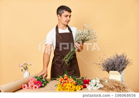 Creative floral design. Colorful plant collection. Expert floral arrangements. Young man florist making bouquet in flower shop standing isolated over beige background 106068656