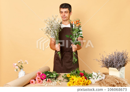 Concentrated serious young male in brown apron working in flower shop standing isolated over beige background being in creating bouquet process choosing plants. Concentrated serious young male in brown apron working in flower shop standing isolated over beige background being in creating bouquet process choosing plants. 106068673