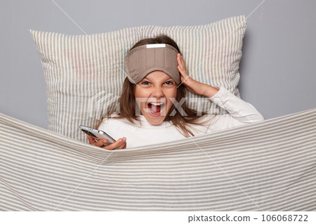 Crazy emotional excited cute girl wearing sleep eye mask resting on pillow under blanket isolated over gray background screaming with amazement 106068722