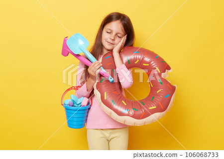 Sleepy little girl holding sandbox toys, bucket and rubber ring isolated over yellow background feeling exhausted after long hours playing at beach with rake and scoop. Sleepy little girl holding sandbox toys, bucket and rubber ring isolated over yellow background feeling exhausted after long hours playing at beach with rake and scoop. 106068733