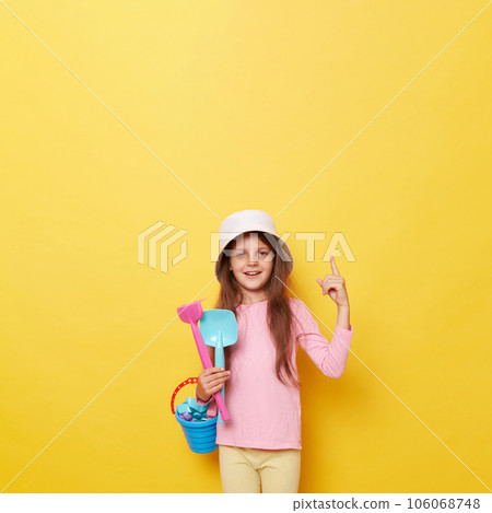 Smiling satisfied little cute girl in panama holding beach sandbox toys rake and shovel isolated over yellow background pointing up showing mockup for promotional text. Smiling satisfied little cute girl in panama holding beach sandbox toys rake and shovel isolated over yellow background pointing up showing mockup for promotional text. 106068748