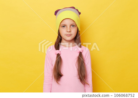 Serious confident little girl with ponytails wearing pink shirt and beanie hat posing isolated over yellow background looking at camera with displeased expression. 106068752