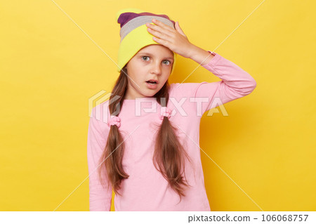 Despair shocked little girl with ponytails wearing pink shirt and beanie hat standing isolated over yellow background having troubles showing facepalm gesture. 106068757