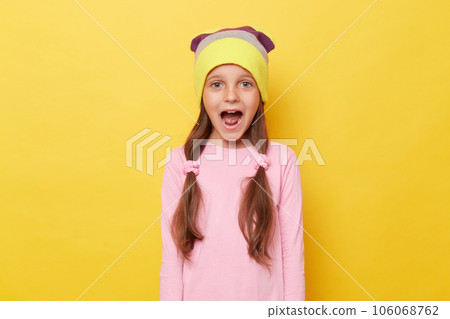 Extremely happy amazed little girl with ponytails wearing beanie hat standing isolated over yellow background screaming with widely open mouth. Extremely happy amazed little girl with ponytails wearing beanie hat standing isolated over yellow background screaming with widely open mouth. 106068762