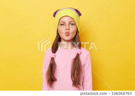 Charming little girl with ponytails wearing beanie hat standing isolated over yellow background sending air kisses keeping lips pout expressing love. 106068763