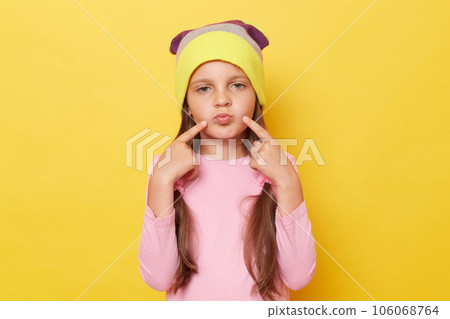 Funny little girl with ponytails wearing pink shirt and beanie hat standing isolated over yellow background pointing at her pout lips. Funny little girl with ponytails wearing pink shirt and beanie hat standing isolated over yellow background pointing at her pout lips. 106068764