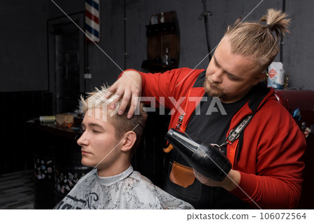 Stylish handsome man barber professional dries the hair of a guy's client with a hair dryer during the haircut process in a hairdresser, or salon 106072564