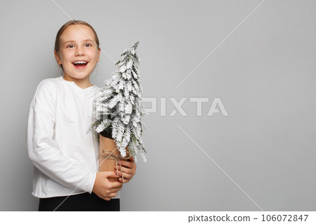 Happy excited emotional child girl holding Christmas tree with snow on light gray banner background Happy excited emotional child girl holding Christmas tree with snow on light gray banner background 106072847
