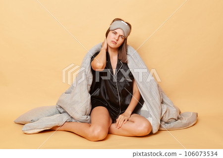 Tired exhausted woman sitting on floor wrapped in plaid wearing pajama, sleep eye mask isolated over beige background feeling pain in neck after uncomfortable sleep. Tired exhausted woman sitting on floor wrapped in plaid wearing pajama, sleep eye mask isolated over beige background feeling pain in neck after uncomfortable sleep. 106073534