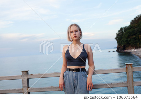 Young blonde woman in tank top standing on the background of blue sea, green coast and cloudy sky 106073564