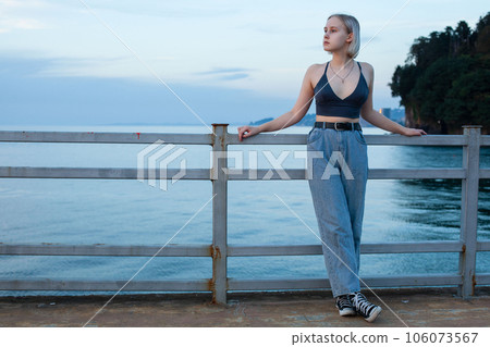 Young woman in jeans and blue tank top standing on pier on the background of blue sea 106073567