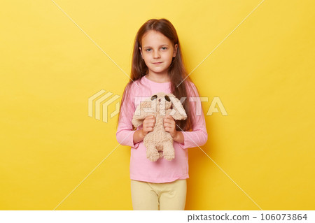 Portrait of adorable little caucasian girl holding fluffy toy in hands standing isolated over yellow background, looking at camera, wearing casual clothing. Portrait of adorable little caucasian girl holding fluffy toy in hands standing isolated over yellow background, looking at camera, wearing casual clothing. 106073864