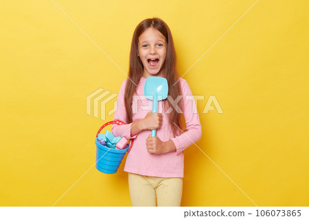 Excited amazed screaming little girl wearing casual clothing holding sandbox toys and bucket isolated over yellow background rejoicing  her playing outdoor. 106073865