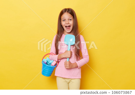 Sunny summer playtime. little cute girl with dark hair holding beach sandbox toys rake shovel and colorful beach bucket. isolated over yellow background screaming with exitement. Sunny summer playtime. little cute girl with dark hair holding beach sandbox toys rake shovel and colorful beach bucket. isolated over yellow background screaming with exitement. 106073867