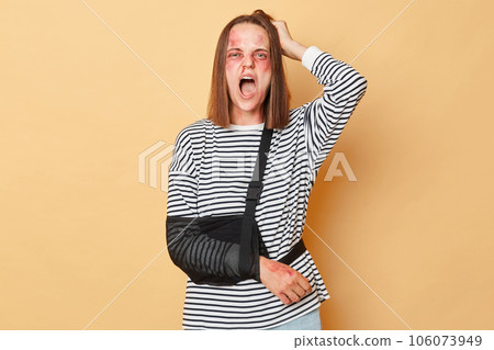 Despair sick woman with scratches and bruises on her face and bandage on arm standing isolated over beige background pulling her hair screaming violence. 106073949