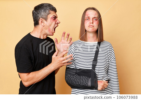 Home violence. Relationship arguingg. Aggressive man screaming with hate and anger, beating his woman, wife with bruise and bandage isolated over beige background. Home violence. Relationship arguingg. Aggressive man screaming with hate and anger, beating his woman, wife with bruise and bandage isolated over beige background. 106073988