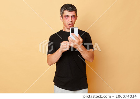 Traumatized beaten man in black T-shirt with bruises and abrasions on his face isolated over beige background using mobile phine chatting with psychologist calling police. Traumatized beaten man in black T-shirt with bruises and abrasions on his face isolated over beige background using mobile phine chatting with psychologist calling police. 106074034