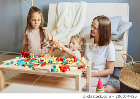 Babysitter's creative playtime. Learning through play and toys. Motherhood and family relationships. Smiling mother playing with little daughters engaged in game activity at home interior 106074450