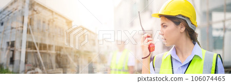 Young attractive construction woman in safety vest with yellow helmet working with radio, standing on building construction site. Home building project. Young attractive construction woman in safety vest with yellow helmet working with radio, standing on building construction site. Home building project. 106074465
