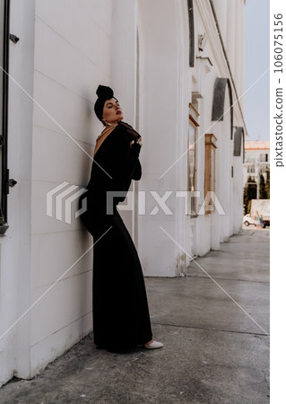 Stylish woman in the city. Fashion photo of a beautiful model in an elegant black dress posing against the backdrop of a building on a city street 106075156