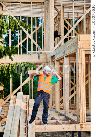 Carpenter building wooden-framed house near the forest. Man holding a large beam on his shoulder, dressed in work clothes and helmet. Idea of modern and sustainable construction. 106076630