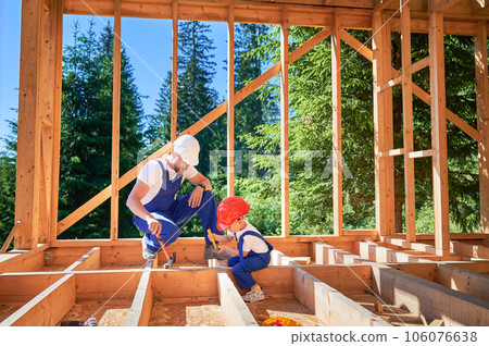 Father with toddler son building wooden frame house. Male worker instructing his son on practice of utilizing hammer to secure nails on construction site. Carpentry and family concept. Father with toddler son building wooden frame house. Male worker instructing his son on practice of utilizing hammer to secure nails on construction site. Carpentry and family concept. 106076638