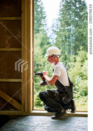 Carpenter constructing wooden framed house. Man worker in glasses working with screwdriver, wearing work overalls and helmet. Concept of modern eco-friendly construction. Carpenter constructing wooden framed house. Man worker in glasses working with screwdriver, wearing work overalls and helmet. Concept of modern eco-friendly construction. 106076639