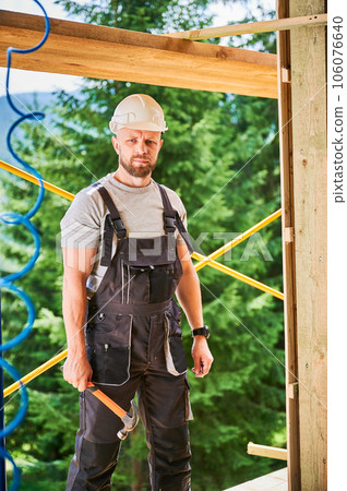 Carpenter constructing wooden frame two-story house near the forest. Portrait of bearded man holding hammer, dressed in protective helmet and overalls. Concept of ecological modern construction. Carpenter constructing wooden frame two-story house near the forest. Portrait of bearded man holding hammer, dressed in protective helmet and overalls. Concept of ecological modern construction. 106076640