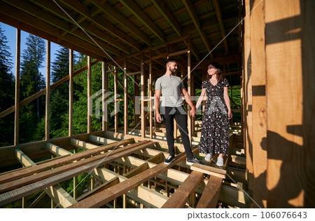 Man and woman inspecting their future wooden frame dwelling nestled near the forest. Youthful couple at construction site in early morning. Concept of contemporary ecological construction. Man and woman inspecting their future wooden frame dwelling nestled near the forest. Youthful couple at construction site in early morning. Concept of contemporary ecological construction. 106076643