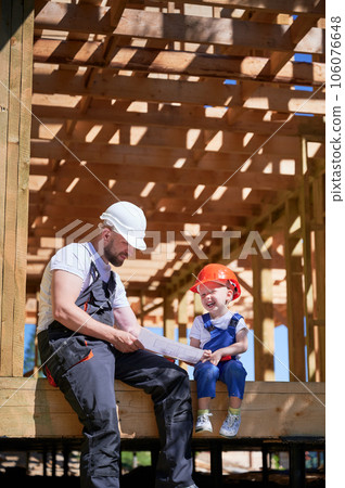 Father with toddler son building wooden frame house. Guys having fun on the edge of balcony, examining the construction plan, wearing helmets and overalls on sunny day. Carpentry and family concept. 106076648