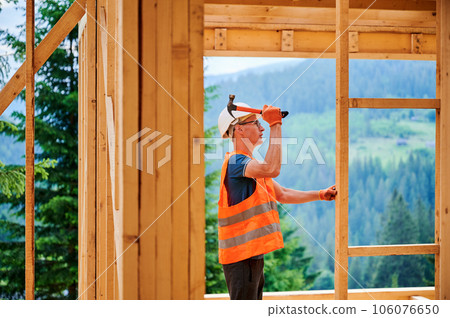 Wooden frame house being built by carpenter. Man wearing glasses hammering nails using hammer and is dressed in a protective helmet and a construction vest. Concept of modern ecological construction. 106076650
