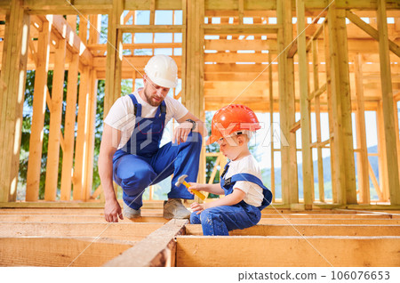 Father with toddler son building wooden frame house. Worker instructing his son on art of hammering nails on construction site, wearing helmets and overalls. Carpentry and family concept 106076653