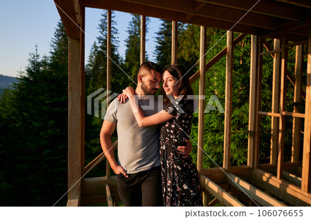 Man and woman inspecting their future wooden frame dwelling nestled in the mountains near forest. Youthful couple at construction site in early morning. Concept of contemporary ecological construction 106076655