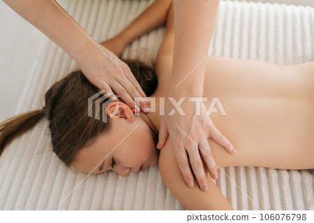 Top view of cute little girl having neck, shoulder and back massage by unrecognizable female masseuse lying on massage table. Adorable preteen kid getting physiotherapy from masseuse for body care. 106076798