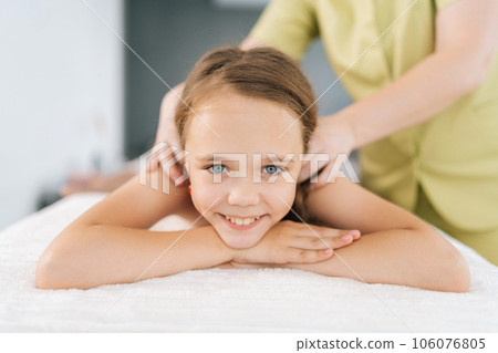 Portrait of adorable little girl having neck, shoulder and back massage by unrecognizable female masseuse at medical clinic, smiling looking at camera. Adorable preteen kid feeling happy and relax. Portrait of adorable little girl having neck, shoulder and back massage by unrecognizable female masseuse at medical clinic, smiling looking at camera. Adorable preteen kid feeling happy and relax. 106076805
