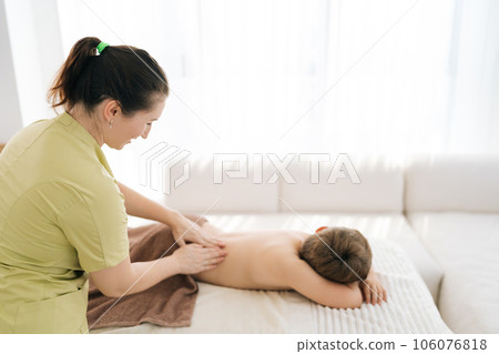 Side view of professional female masseur massaging back of five year old boy in spa salon. Close up of therapeutic, preventive massage session using oil, relaxing muscles, improving blood circulation. Side view of professional female masseur massaging back of five year old boy in spa salon. Close up of therapeutic, preventive massage session using oil, relaxing muscles, improving blood circulation. 106076818