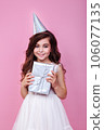 Smiling kid girl in birthday party princess hat holding gift box on a pink background 106077135