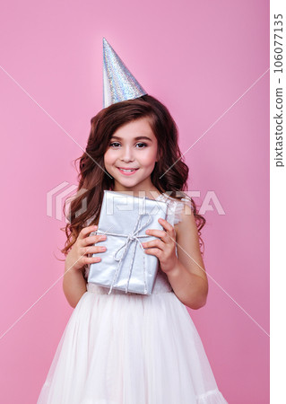 Smiling kid girl in birthday party princess hat holding gift box on a pink background Smiling kid girl in birthday party princess hat holding gift box on a pink background 106077135