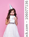 Portrait of surprised kid girl n birthday party princess hat holding gift box on a pink studio background 106077136