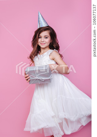 Smiling kid girl in birthday party princess hat holding gift box on a pink background. Kid birthday Smiling kid girl in birthday party princess hat holding gift box on a pink background. Kid birthday 106077137