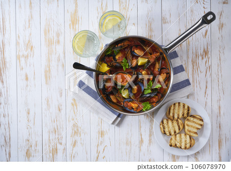 Steamed mussels in shells and tomato sauce with roasted bread and greens on a white table, top view. Steamed mussels in shells and tomato sauce with roasted bread and greens on a white table, top view. 106078970