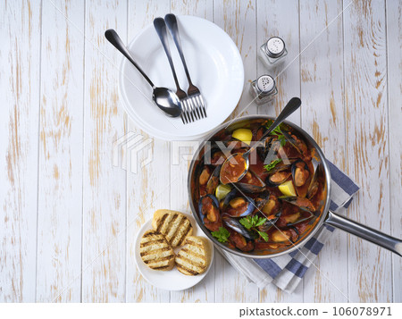 Steamed mussels in shells and tomato sauce with roasted bread and greens on a white table. Seafood dinner. Steamed mussels in shells and tomato sauce with roasted bread and greens on a white table. Seafood dinner. 106078971